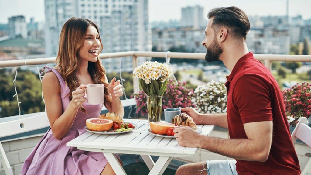 A couple having breakfast on a balcony and smiling and talking.