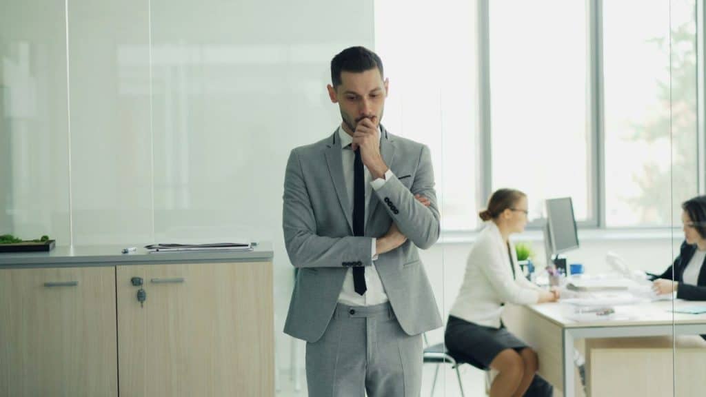 A man in a grey suit stands with his hand on his chin in an office.