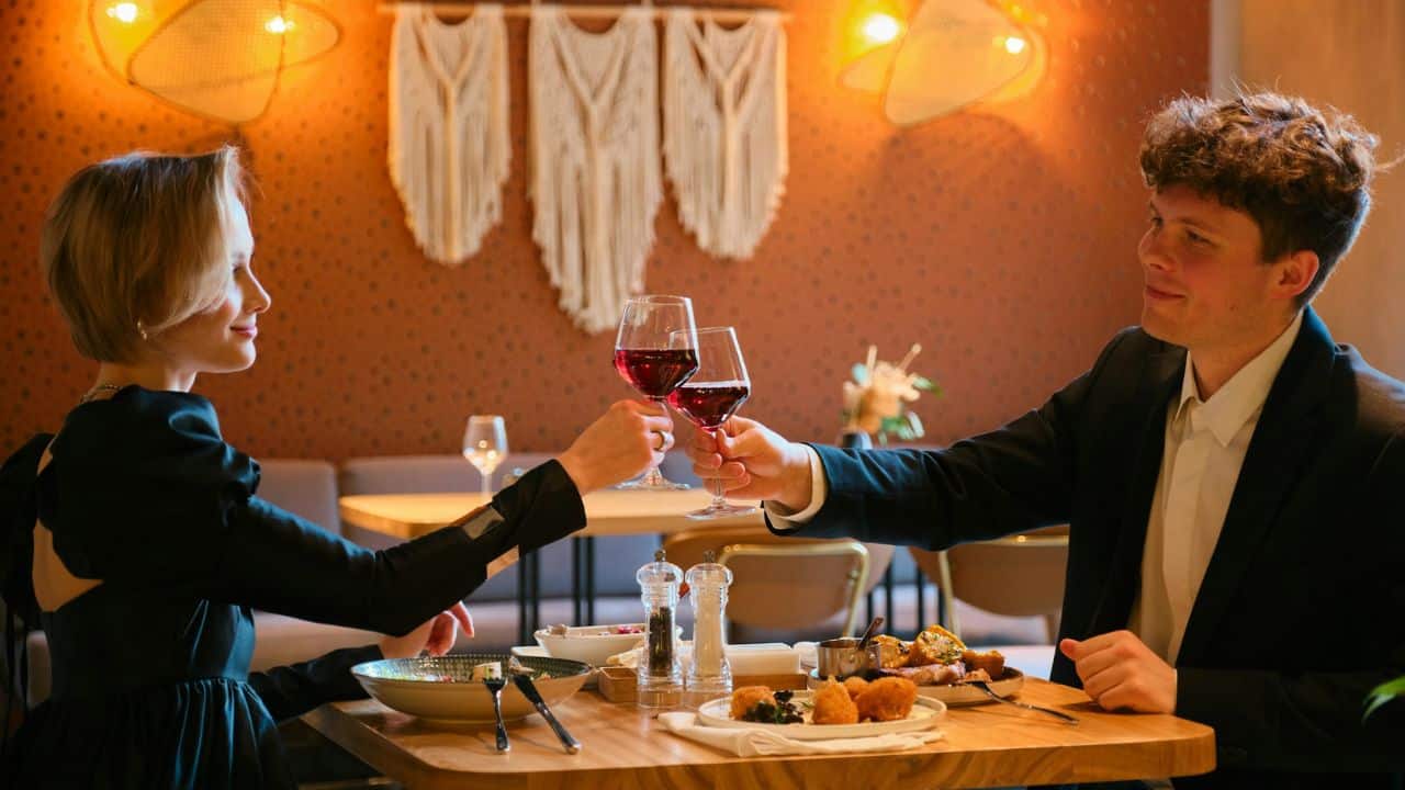 A man and woman toast with glasses of red wine at a restaurant dinner table.