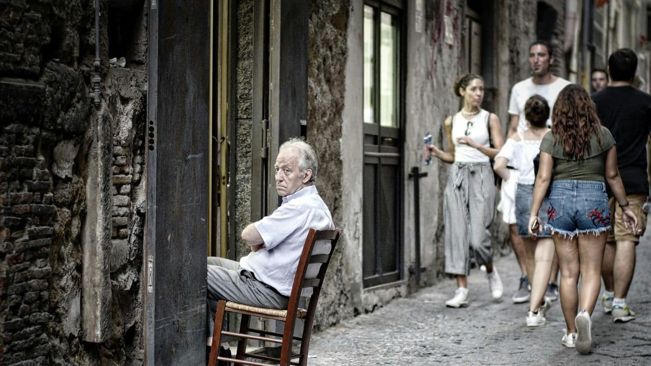 An older man sits in a chair in an alleyway while several younger people walk past.