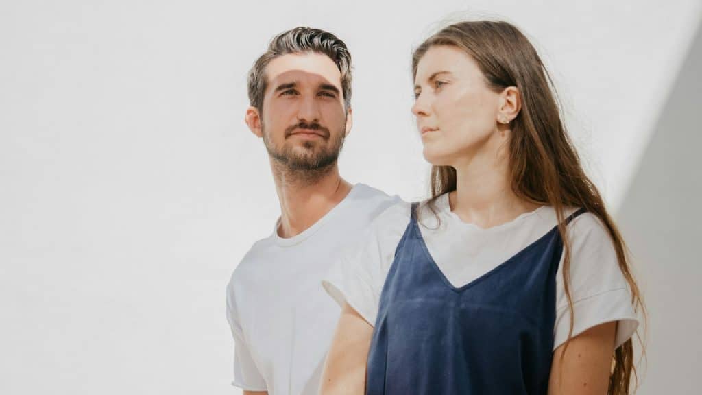 A bearded man and a woman standing side by side and gazing in different directions with solemn expressions.