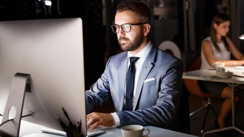 A man in a suit works at a computer at night while a woman sits behind him.