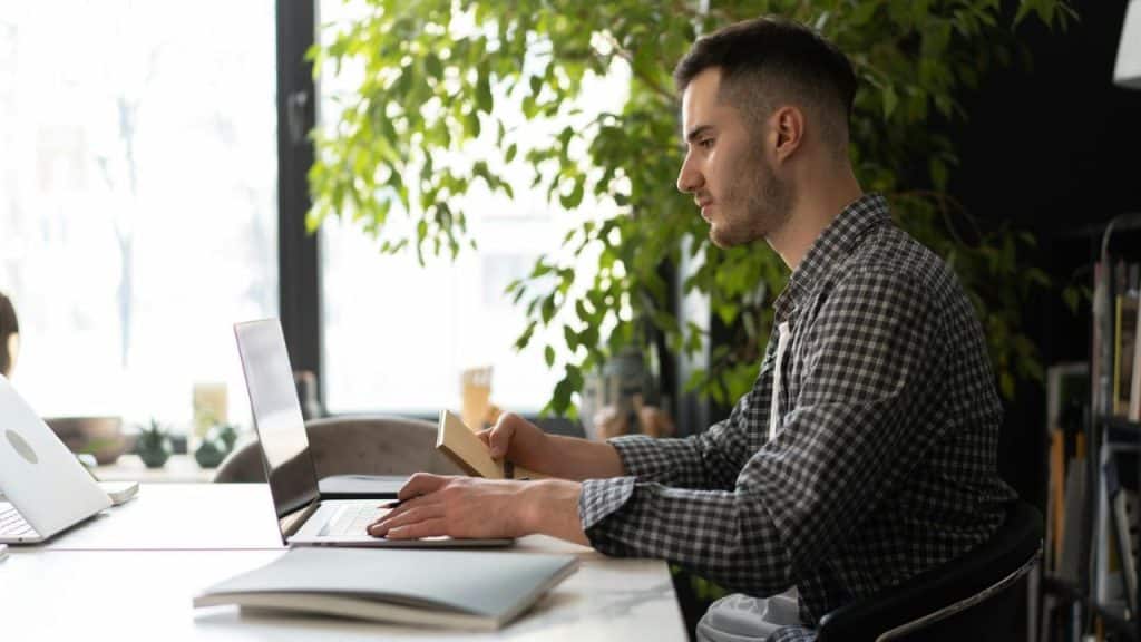 A man in a plaid shirt sits at a desk using a laptop and holding a notebook.