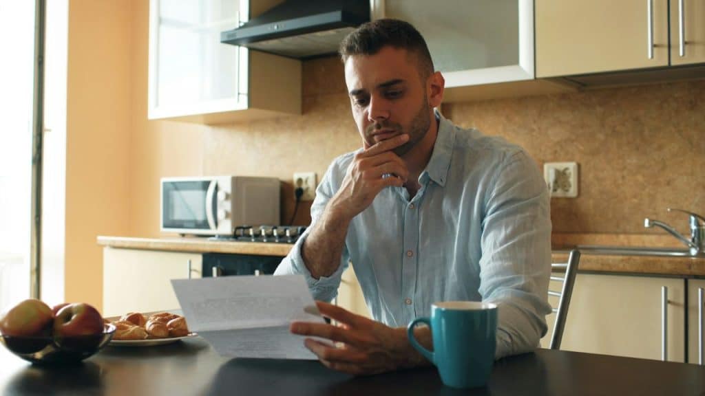 A man sits at a kitchen table with hand on chin, reading a document intently.©Curated Lifestyle/Unsplash.com