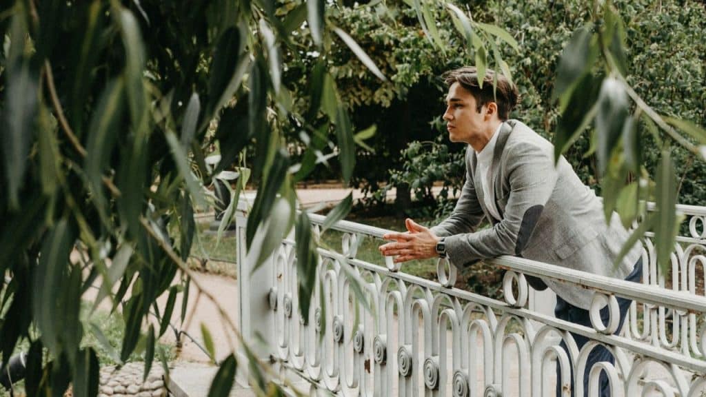 A man in a grey jacket leans against a white metal bridge railing in a park.