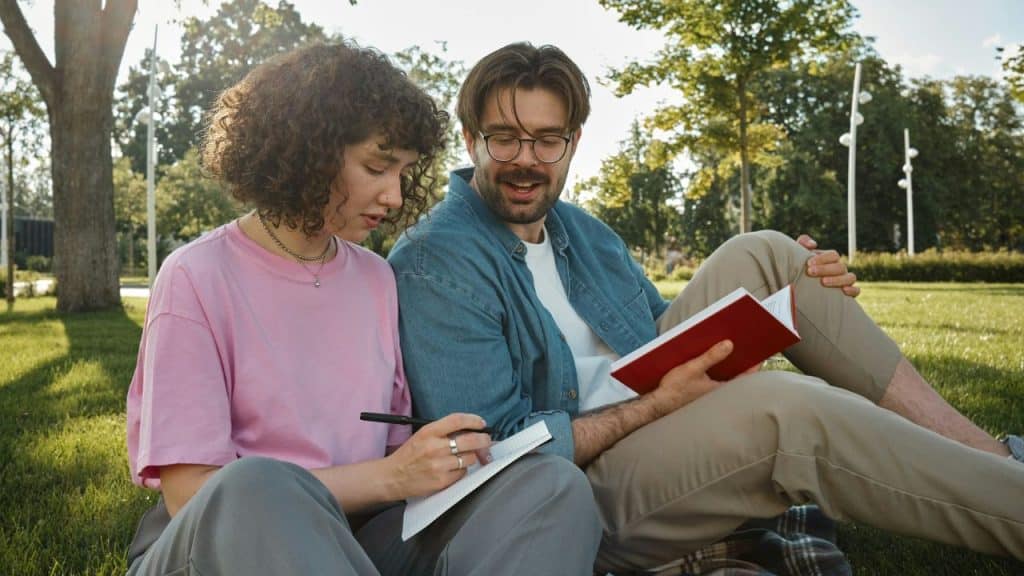 A man and woman sit on grass; she writes in a notebook while he holds a book.