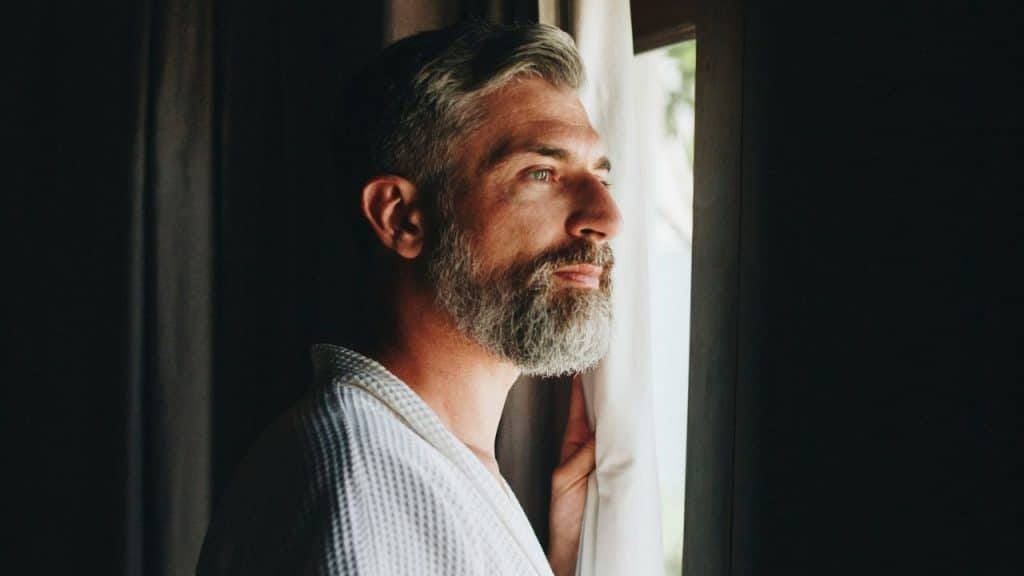 A bearded man in a white robe looks out a window through a gap in curtains.