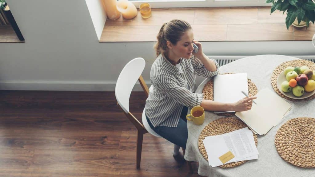 A woman sits at a table on the phone with papers and a laptop.