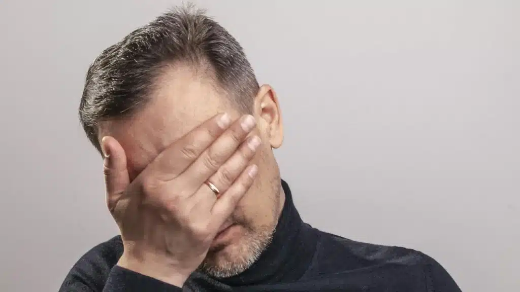 A man in a dark turtleneck covers his eyes with one hand against a plain background.