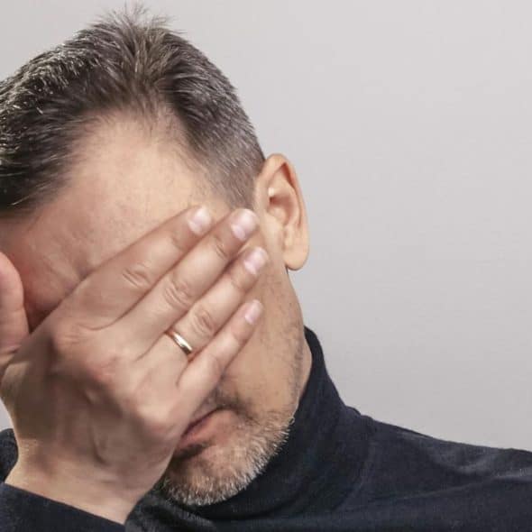 A man in a dark turtleneck covers his eyes with one hand against a plain background.