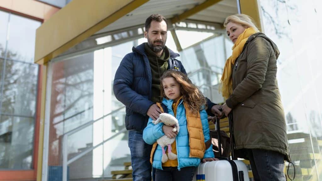 A man and woman stand outdoors near a young child holding a stuffed animal.