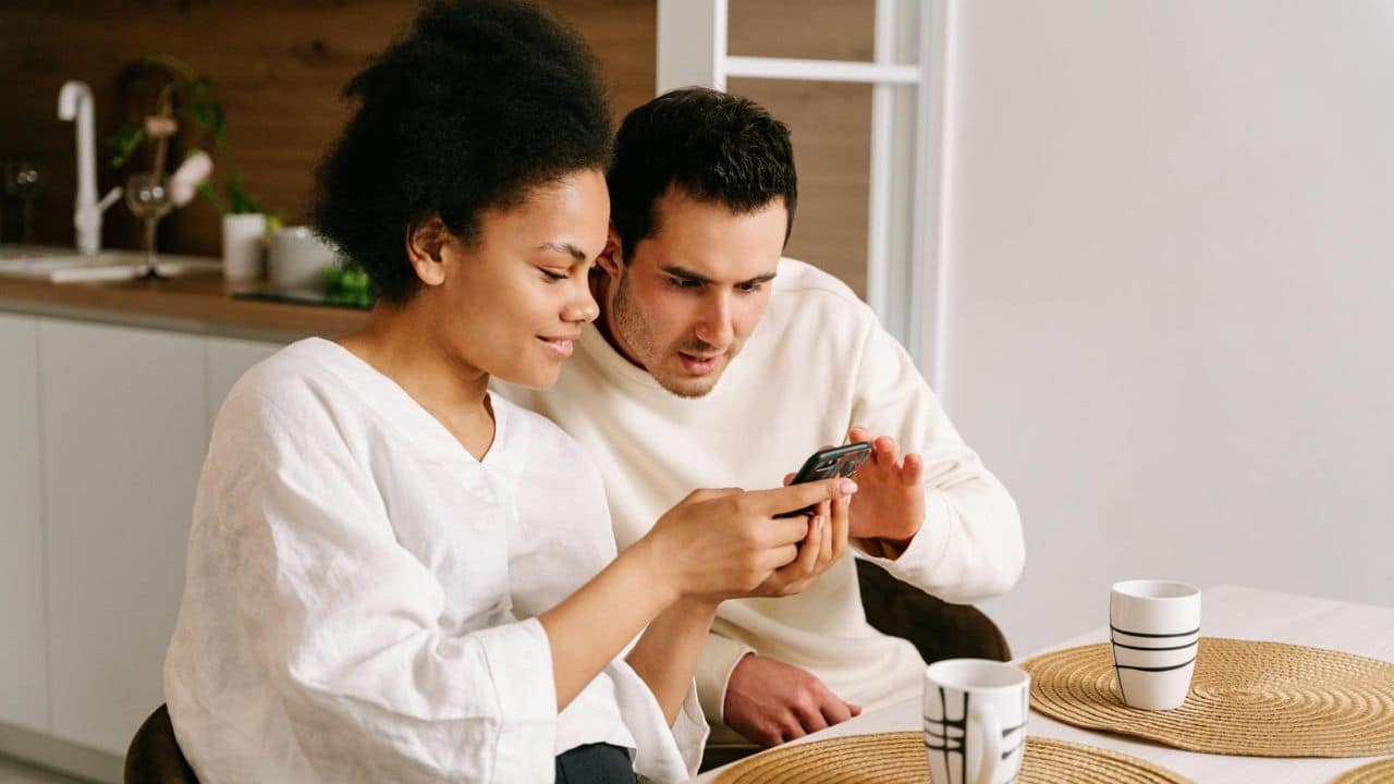 A man and woman sit together at a table, both looking closely at a smartphone.