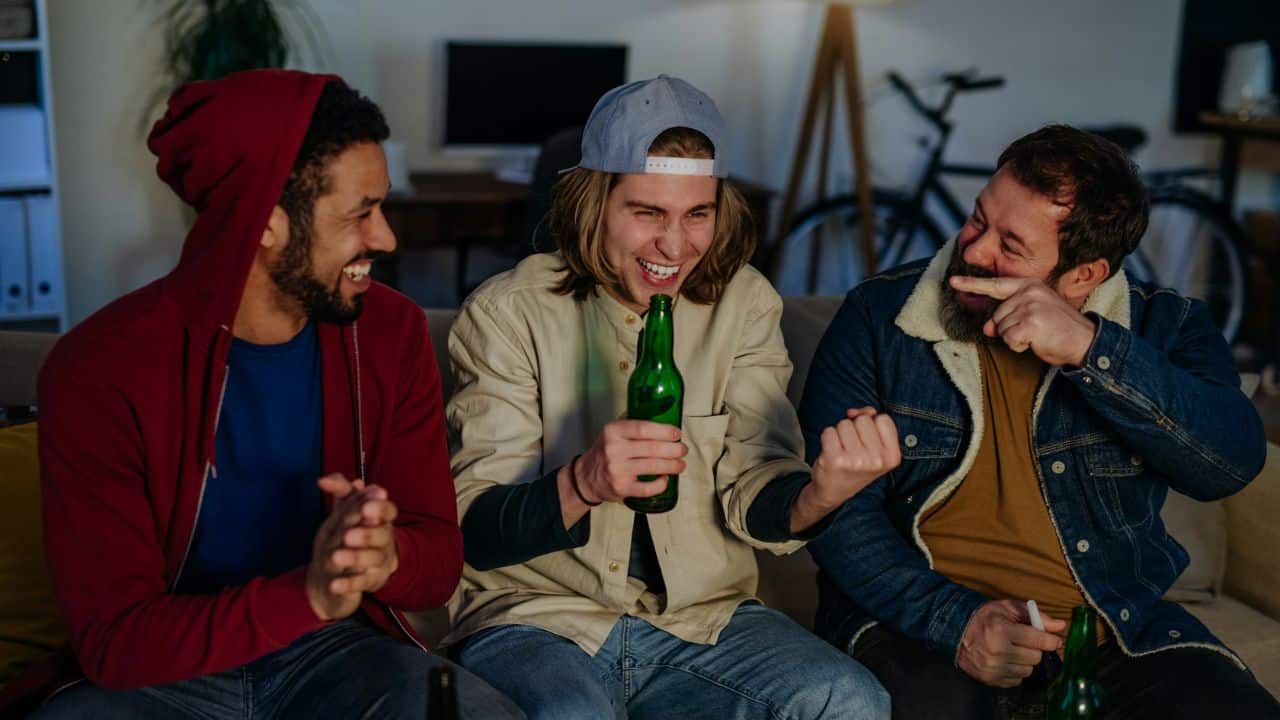 Three men sit together on a sofa, laughing while one holds a green bottle.