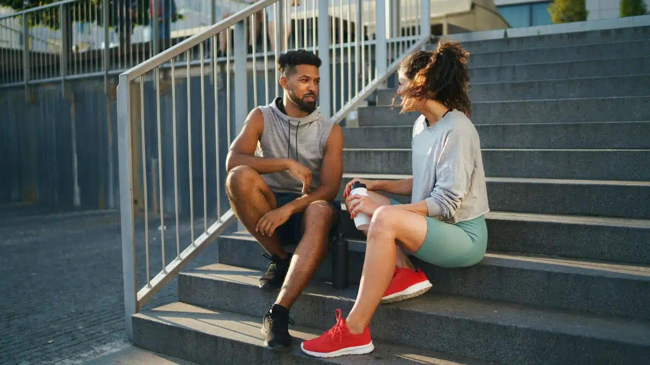 A man and woman in athletic wear sit on concrete stairs and talk outdoors.