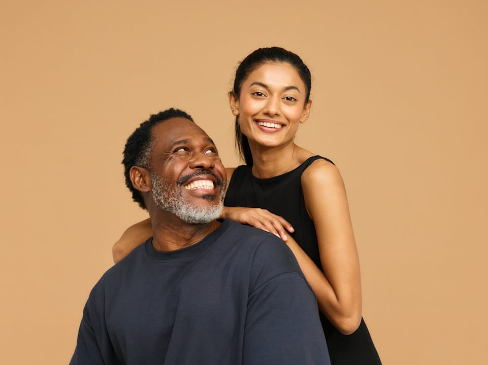 A bearded older man smiling at a younger woman who’s resting her arms on his shoulders.