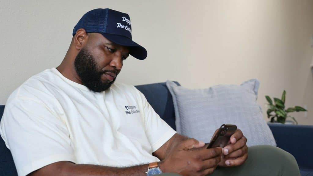A bearded man wearing a baseball cap sits on a sofa looking down at his smartphone.