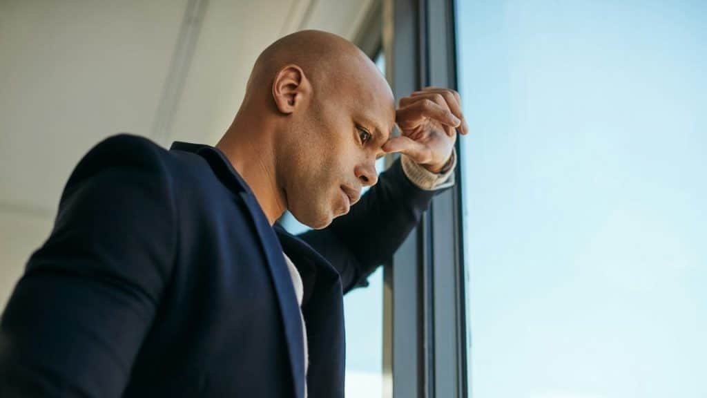 A man in a dark blazer leans against a window with his hand against his forehead.