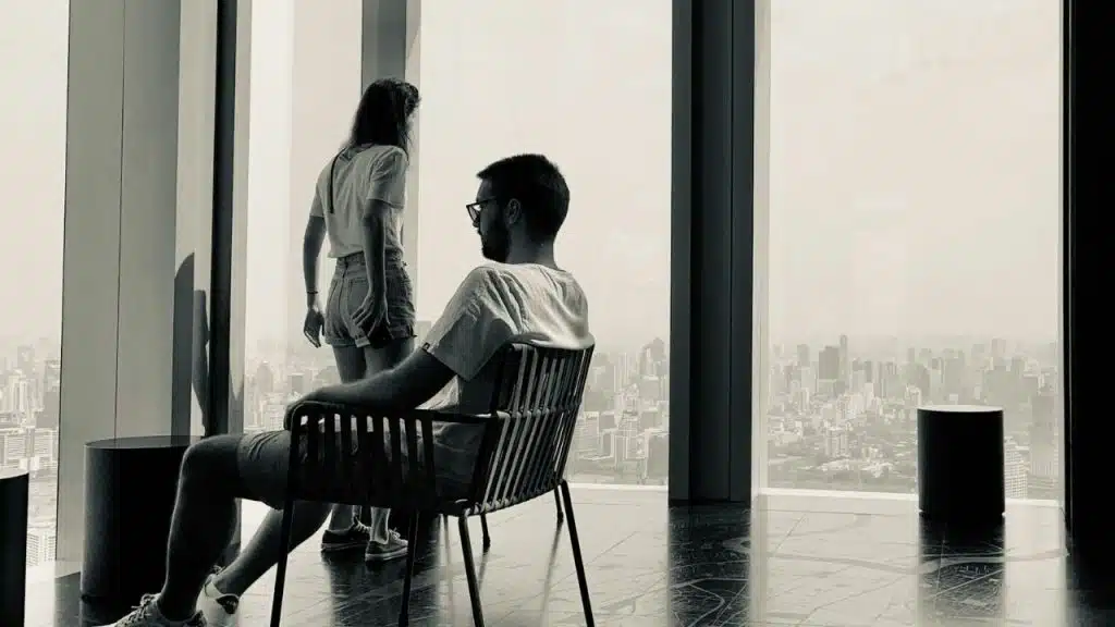 Black and white photo of a man sitting while a woman stands by a window.