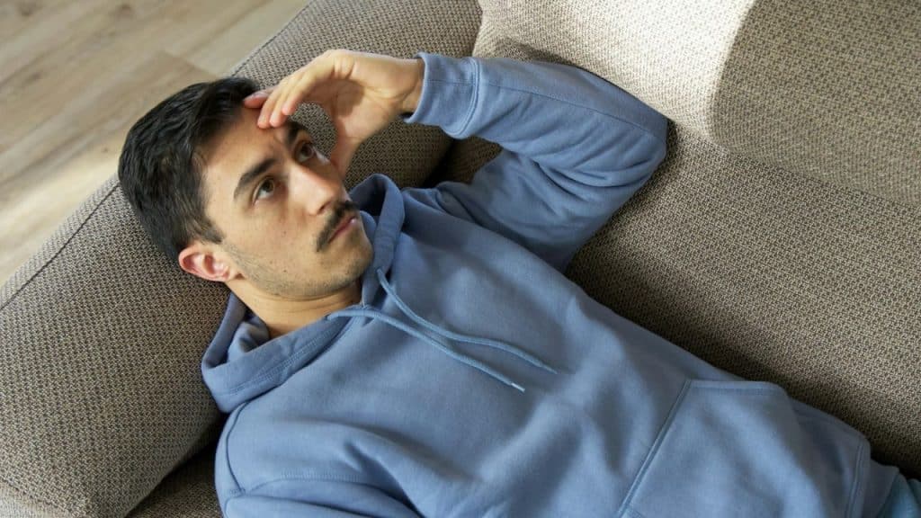 A man with a mustache in a blue hoodie lies on a couch, touching his forehead.