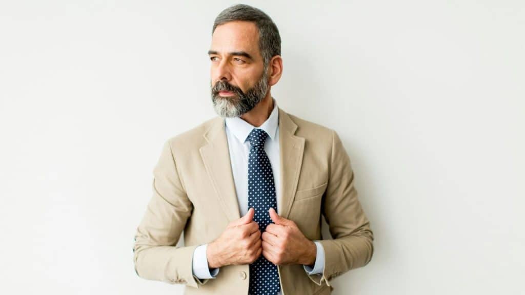 A man with a graying beard and tan suit jacket looks off to the side.