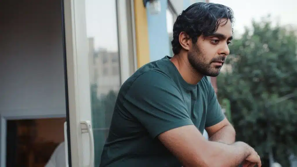 A man in a green t-shirt looks thoughtfully into the distance while leaning on a balcony.