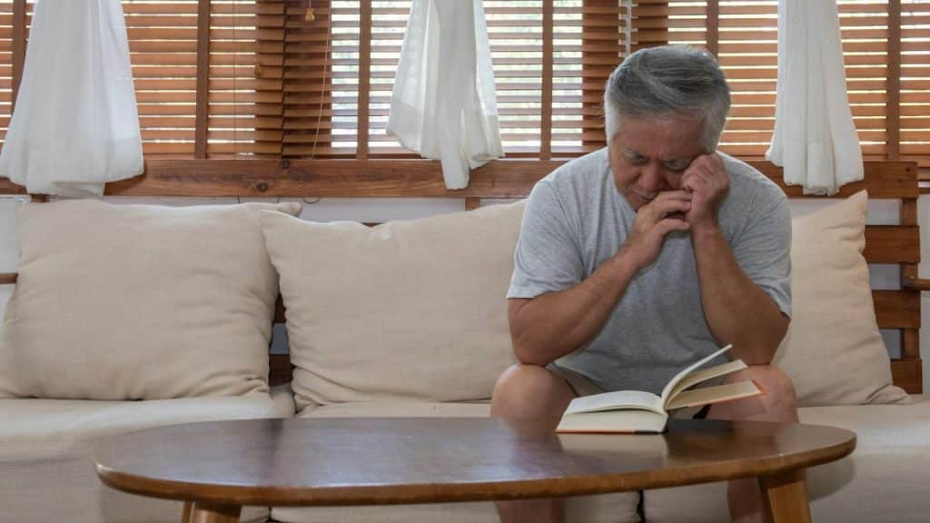 An older man sits on a couch with his head down, appearing distressed near an open book.