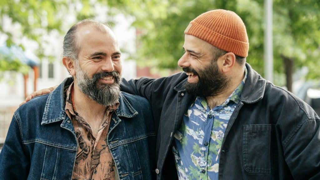 Two men with beards stand close together and smile, with one man's arm around the other.