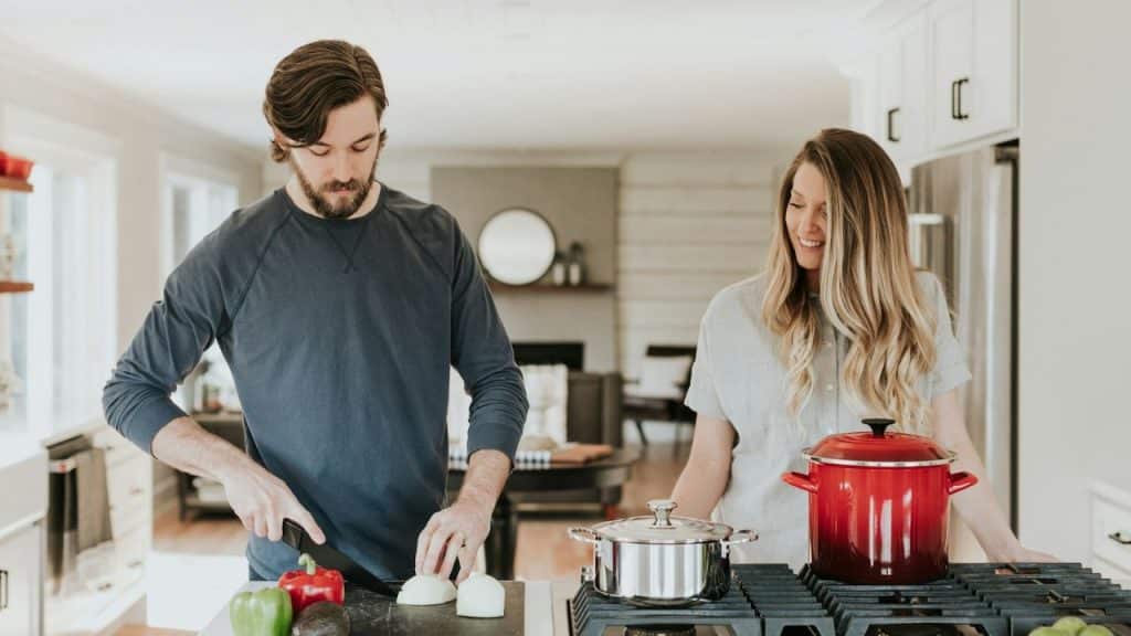 A man chops onions at a kitchen counter while a woman watches him and smiles.