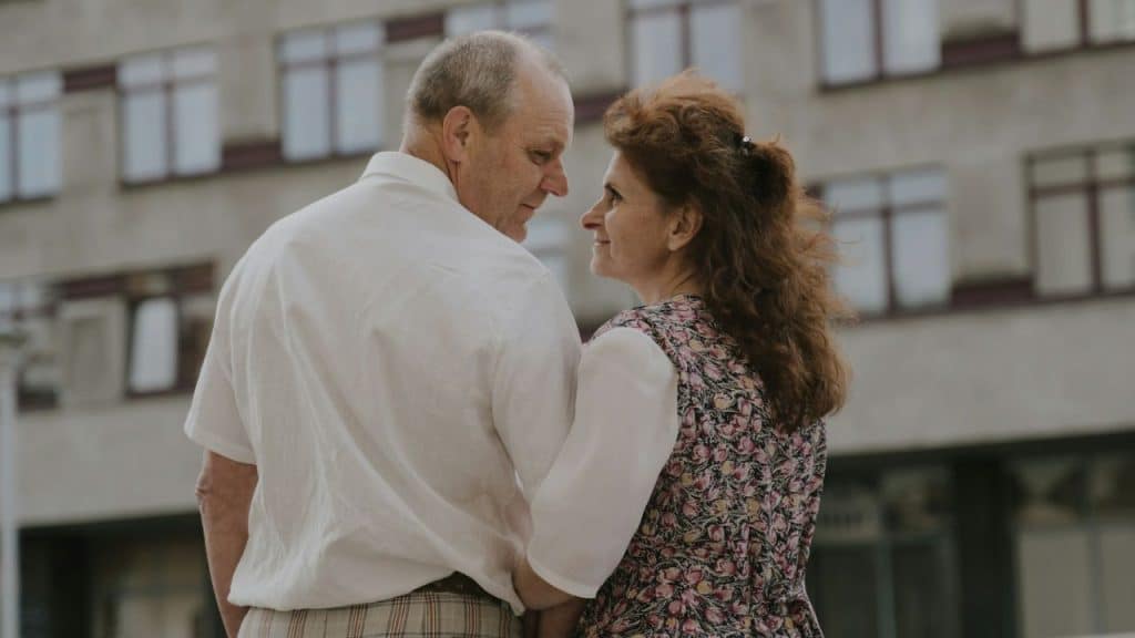 An older couple stands outdoors together, looking into each other's eyes and smiling.