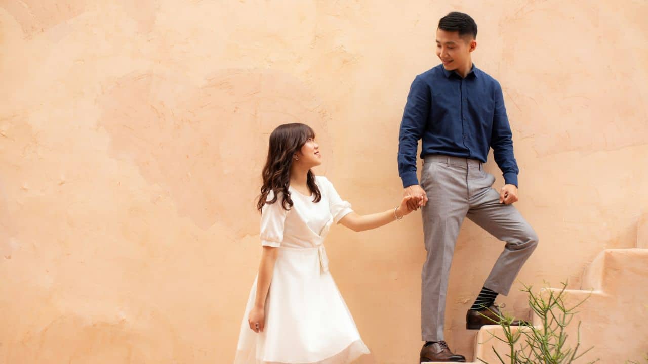 A man and woman hold hands while walking up outdoor steps against a tan wall.
