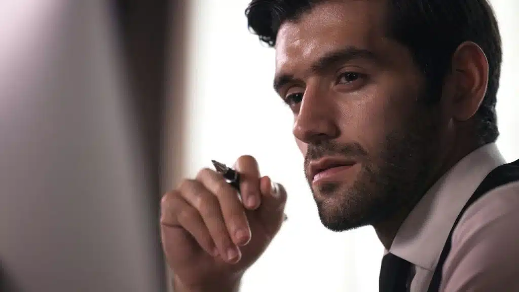 A man in a white dress shirt and black tie holds a pen while looking forward.