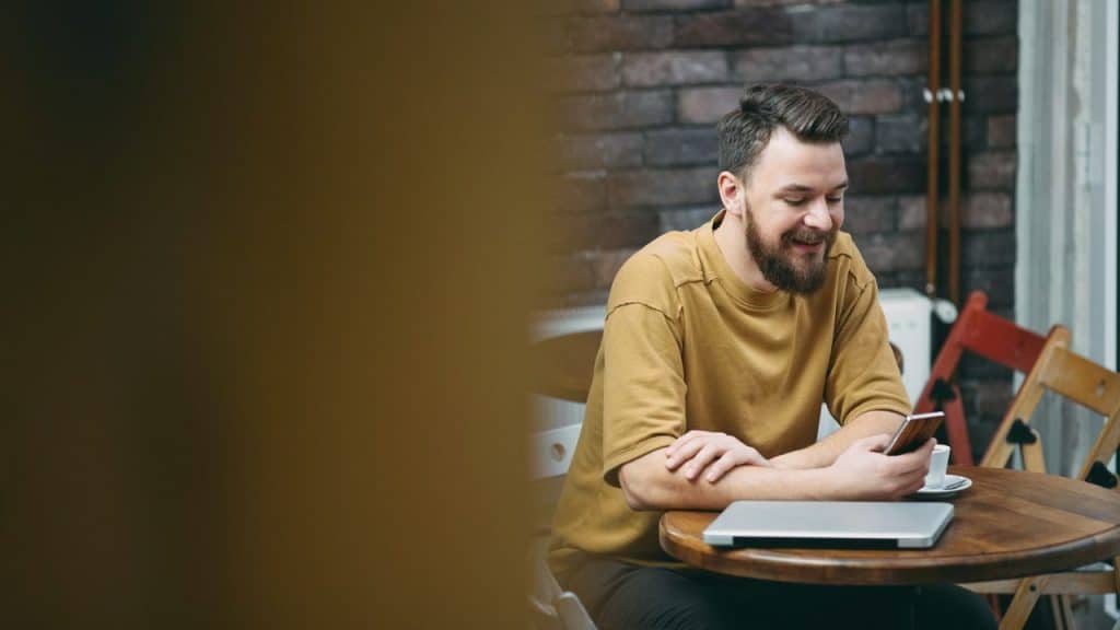 A smiling, bearded man looks at his phone while sitting at a table with a laptop.