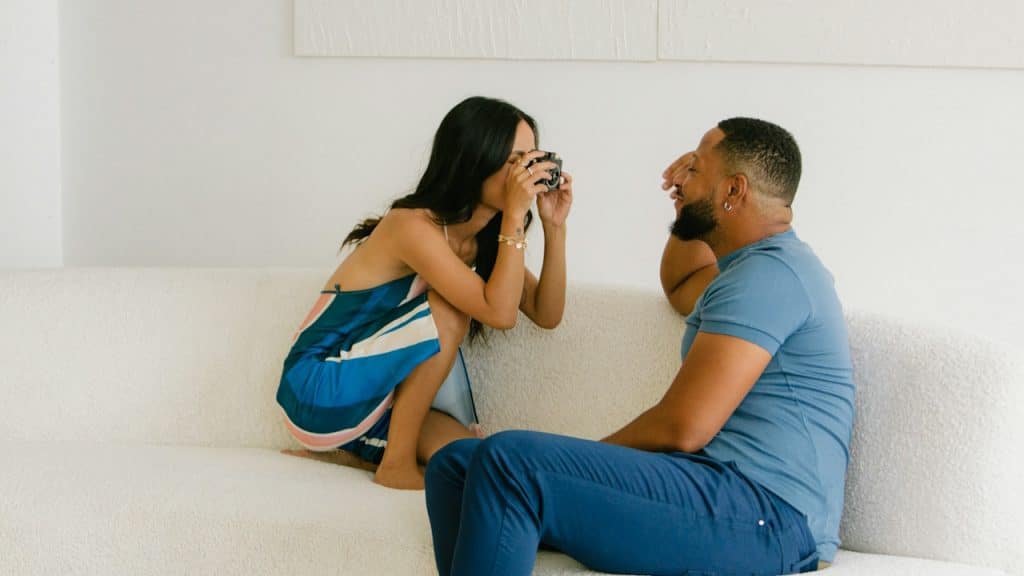 A woman taking a close-up picture of a bearded man while they sit on a couch.