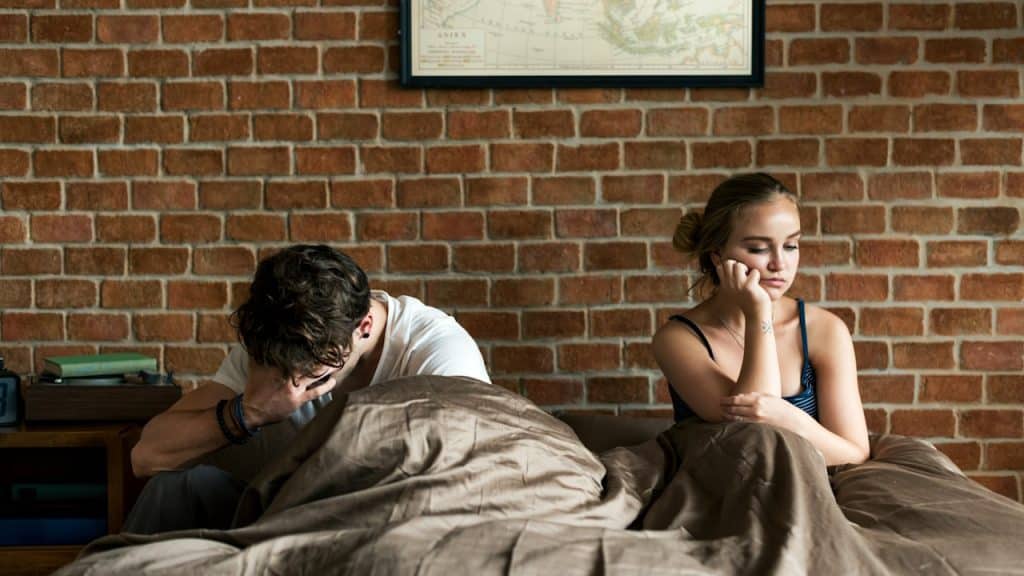 A couple in bed where the man looks upset and the woman looks apathetic and detached.