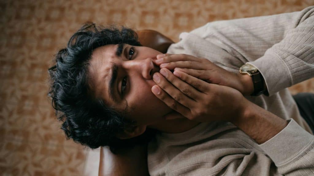 A man lies down with his hands covering his mouth, looking directly at the camera.