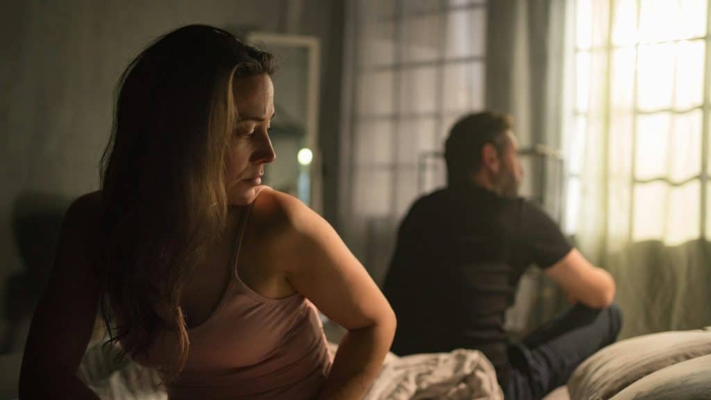 A man and a woman sit on opposite sides of a bed, facing away from each other with somber expressions.