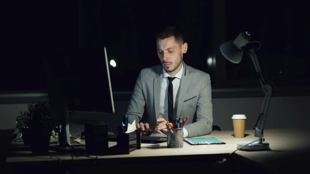 A man in a suit works at a computer in a dark office at night.