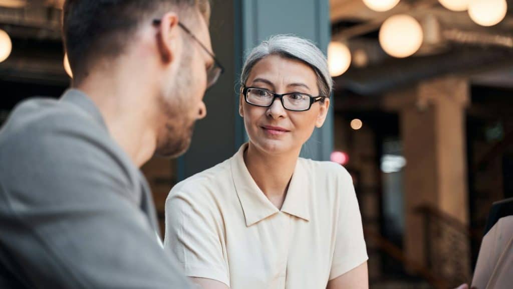 A woman with glasses and grey hair looks at a man in an indoor setting.