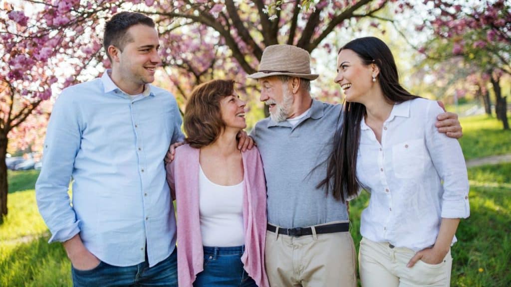 Four people of different ages smiling and walking together in a park with pink blossoms.