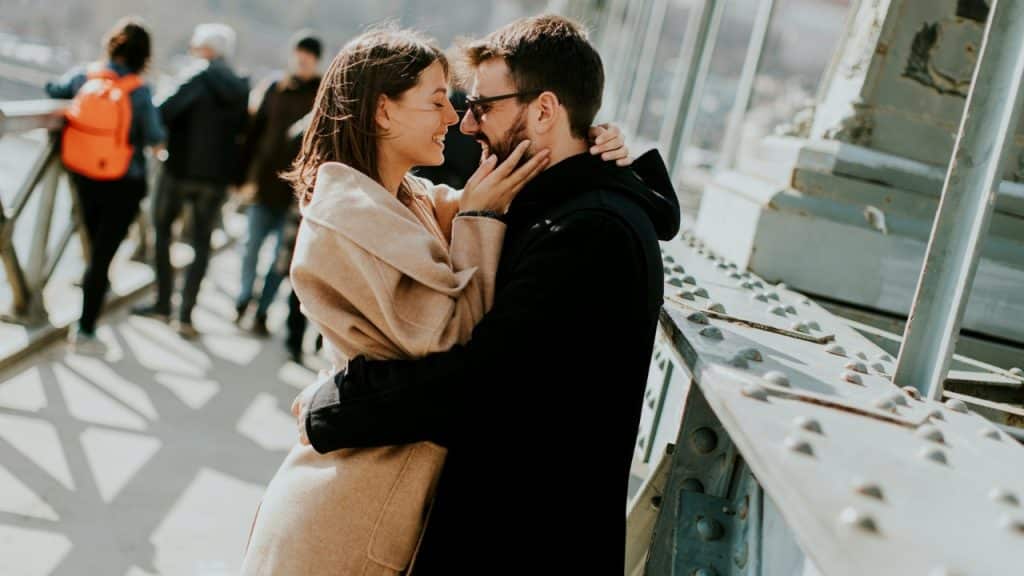 A man and woman in coats embrace while standing on a bridge outdoors in sunlight.