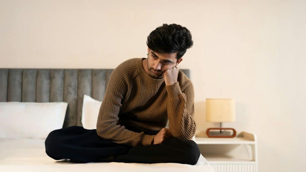 A man in a brown sweater sits cross-legged on a bed, looking down thoughtfully.