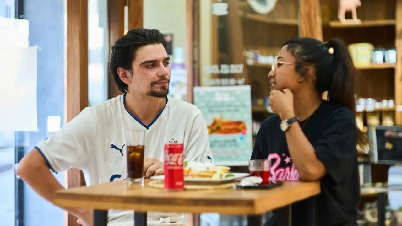 A man and woman sit at an outdoor table with food and drinks talking.