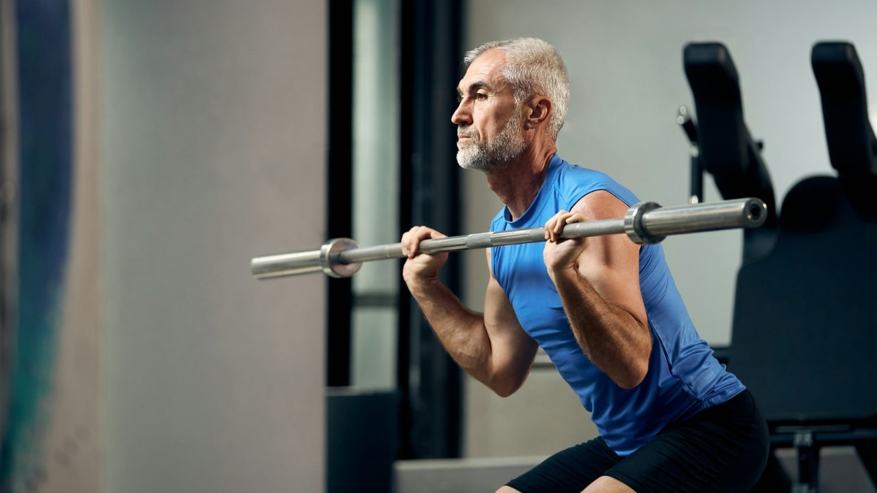 A focused older man in athletic wear performs a front squat with a weighted barbell.