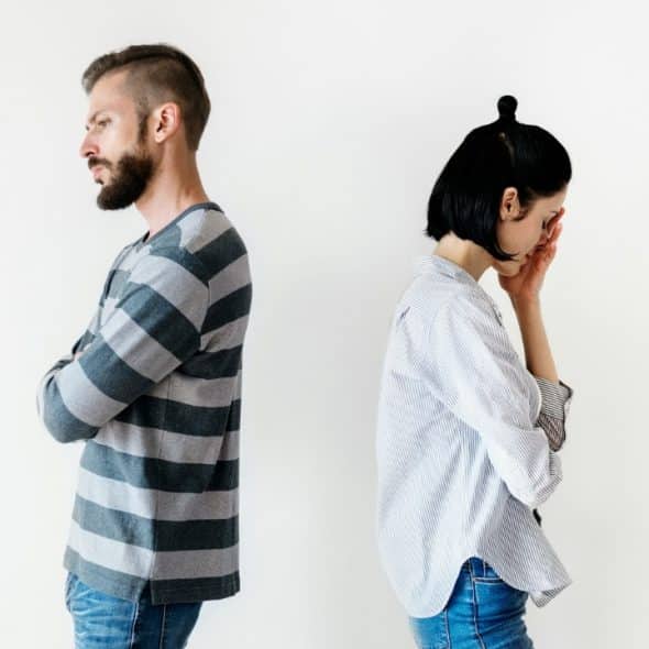 A man and a woman stand back-to-back against a plain white background looking distressed.