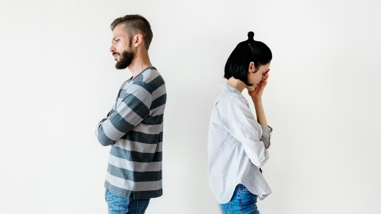 A man and a woman stand back-to-back against a plain white background looking distressed.