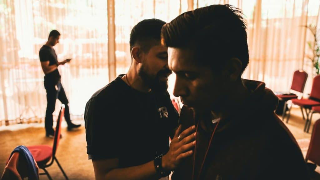 A man stands with his hand on another man's chest in a dimly lit, indoor setting.