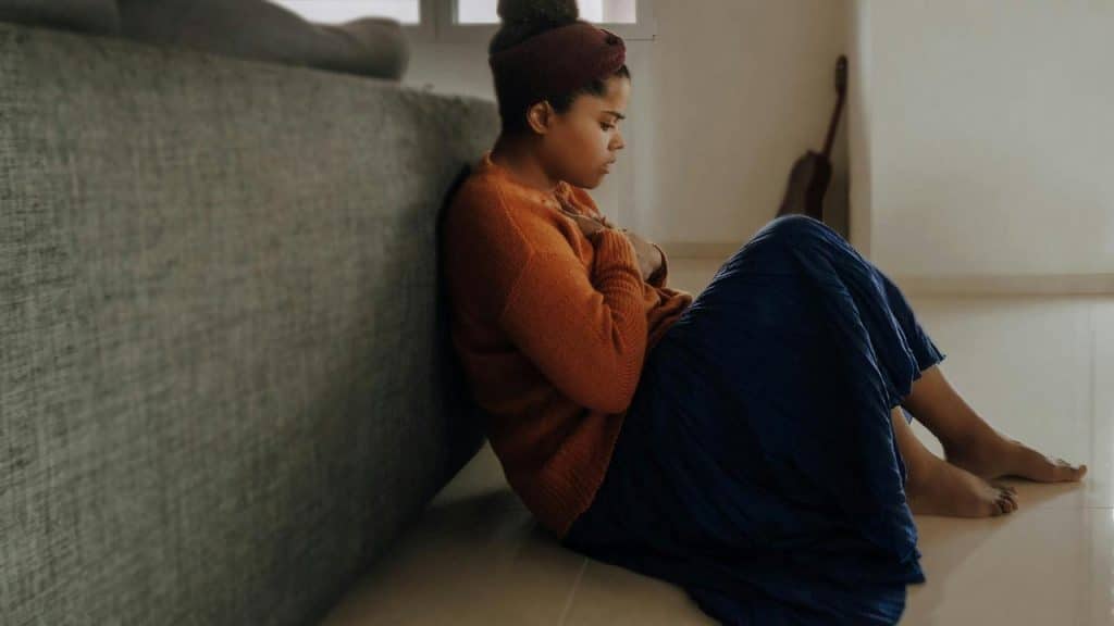 A woman sitting on the floor leaning against a couch, looking down.