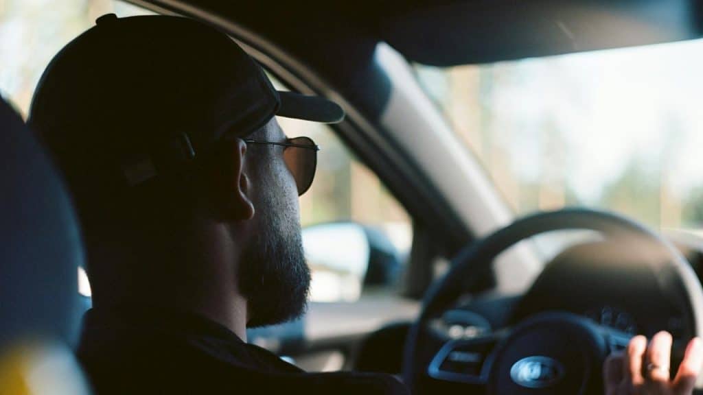 A man wearing a cap and sunglasses driving a car.