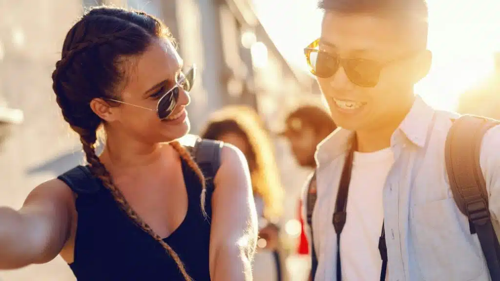 Two smiling people wearing sunglasses and backpacks stand facing each other in the sunlight.