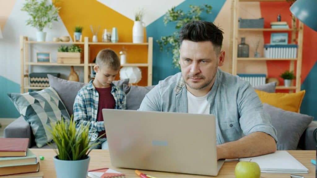 A man works on a laptop while a young boy sits on a couch behind him.